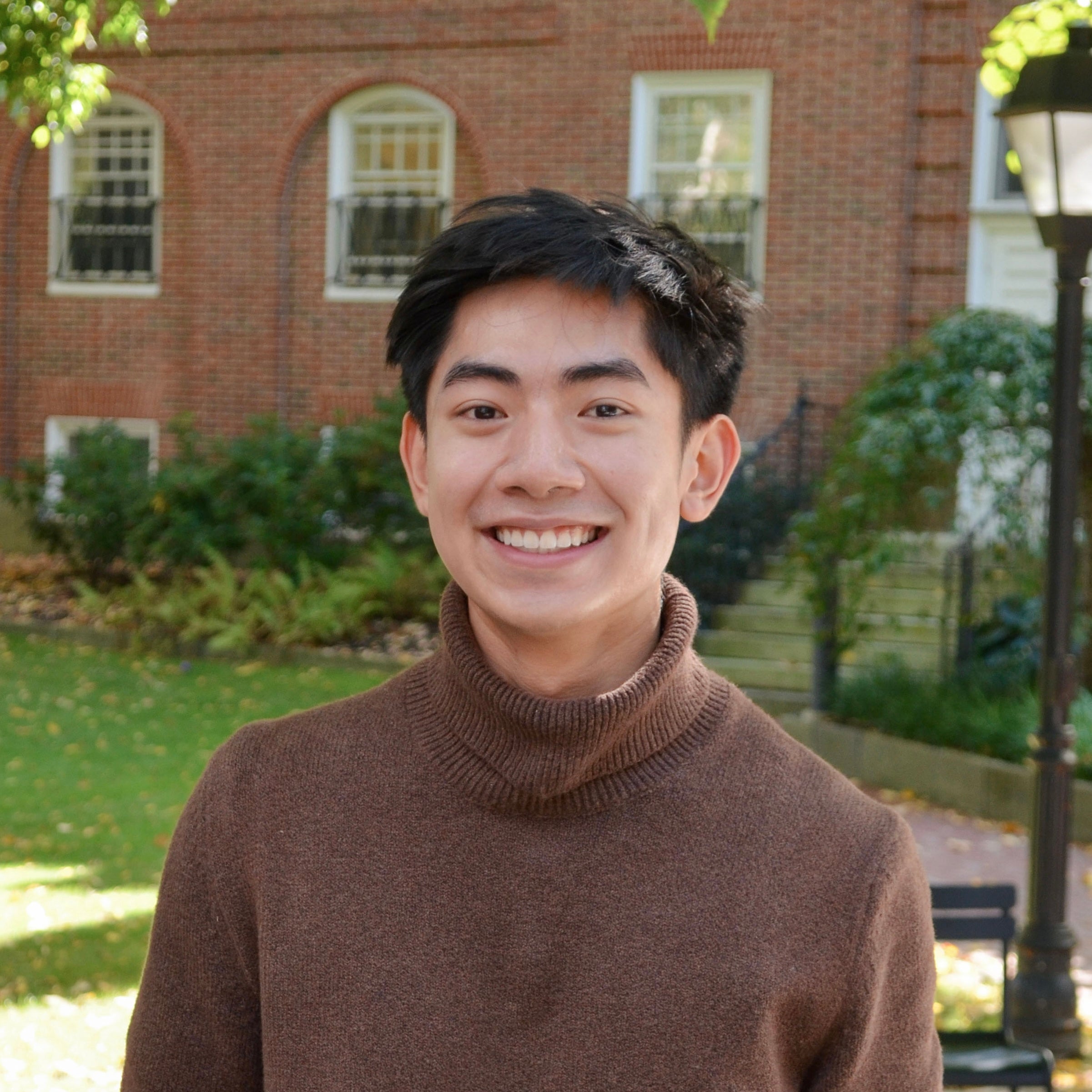 a headshot of Ricky Truong. he is outside, and the background is of a red brick building with windows and green shrubs and grass. Ricky is smiling at the camera and is wearing a brown turtle neck sweater