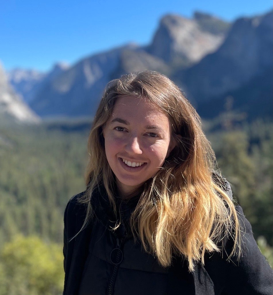a headshot of Tina Nepheli Vartziotis. She has brown-blonde hair just past her shoulders and it is gathered over one shoulder. Her background is blurred, but she is outside with mountains and greenery behind her and a clear blue sky. Tina smiles at the camera and is wearing a black long sleeve shirt.