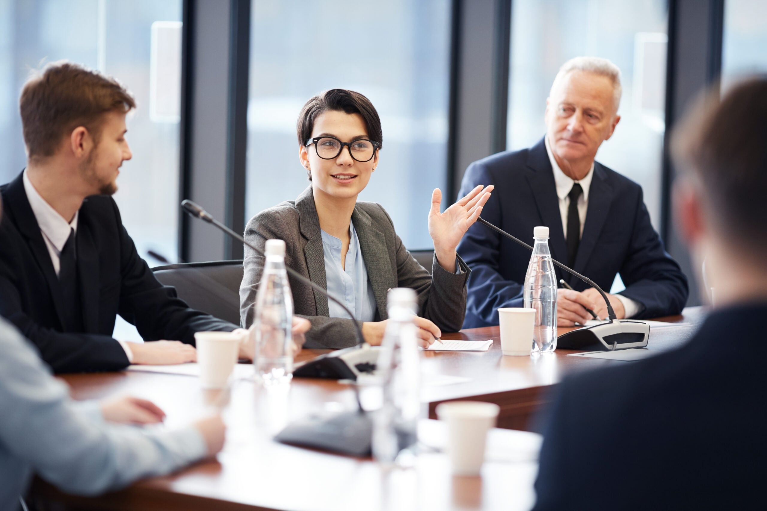 A group of professionals sits around a conference table in a modern office setting. A woman in glasses and a gray blazer is speaking, gesturing with her hand as she addresses the group. Her colleagues, dressed in business attire, listen attentively, with one older man looking at her with interest. Microphones and water bottles are arranged on the table, indicating a formal meeting or discussion. The large windows in the background bring in natural light, creating an open and professional atmosphere.