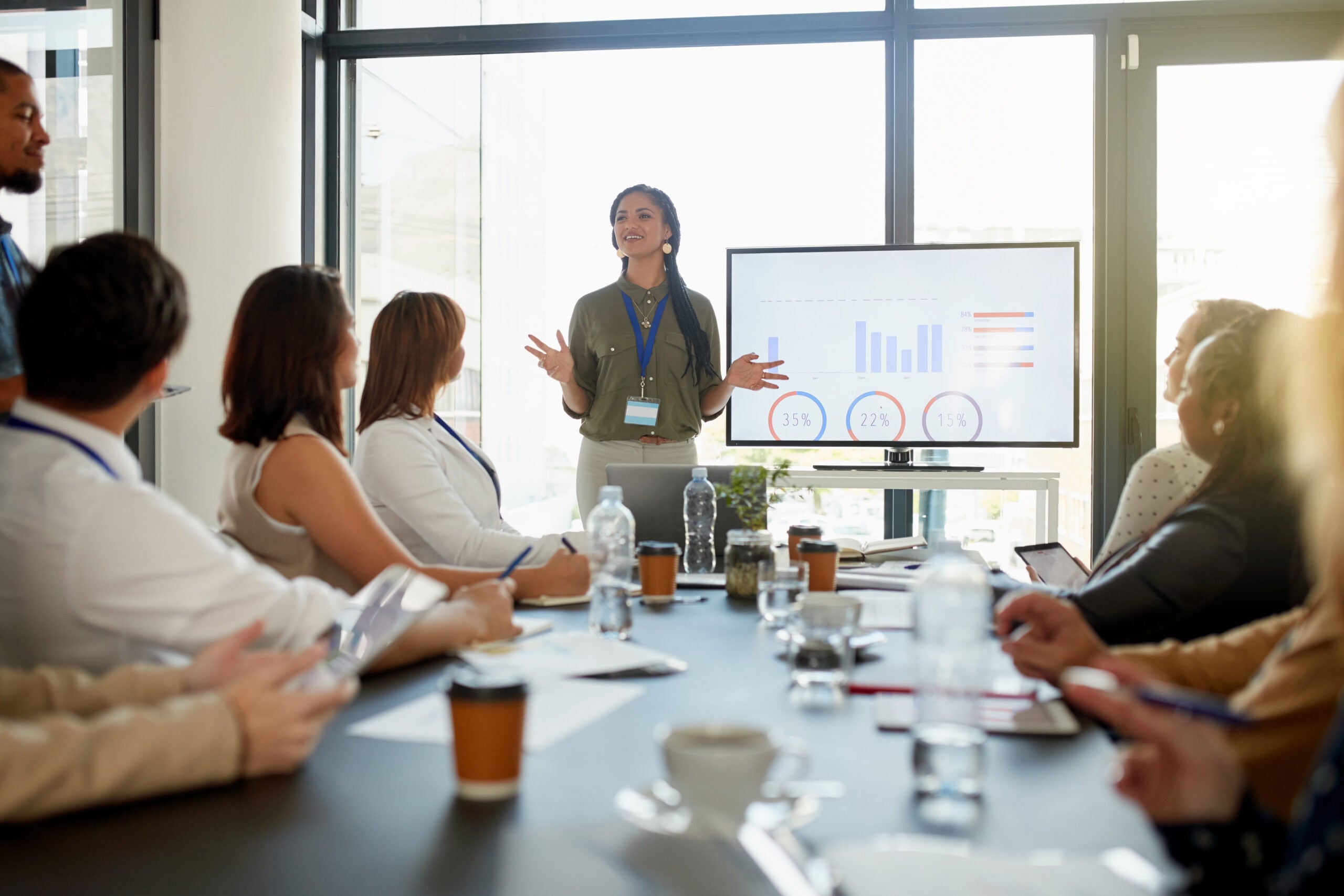 A woman stands at the front of a conference room, presenting to a group seated around a table. She gestures confidently as she explains data displayed on a screen behind her, which shows various charts and percentages. The attendees, wearing conference lanyards, listen attentively, some taking notes. The table is arranged with water bottles, coffee cups, and notepads, suggesting an organized, professional setting. Large windows in the background bring in natural light, enhancing the open and collaborative atmosphere.