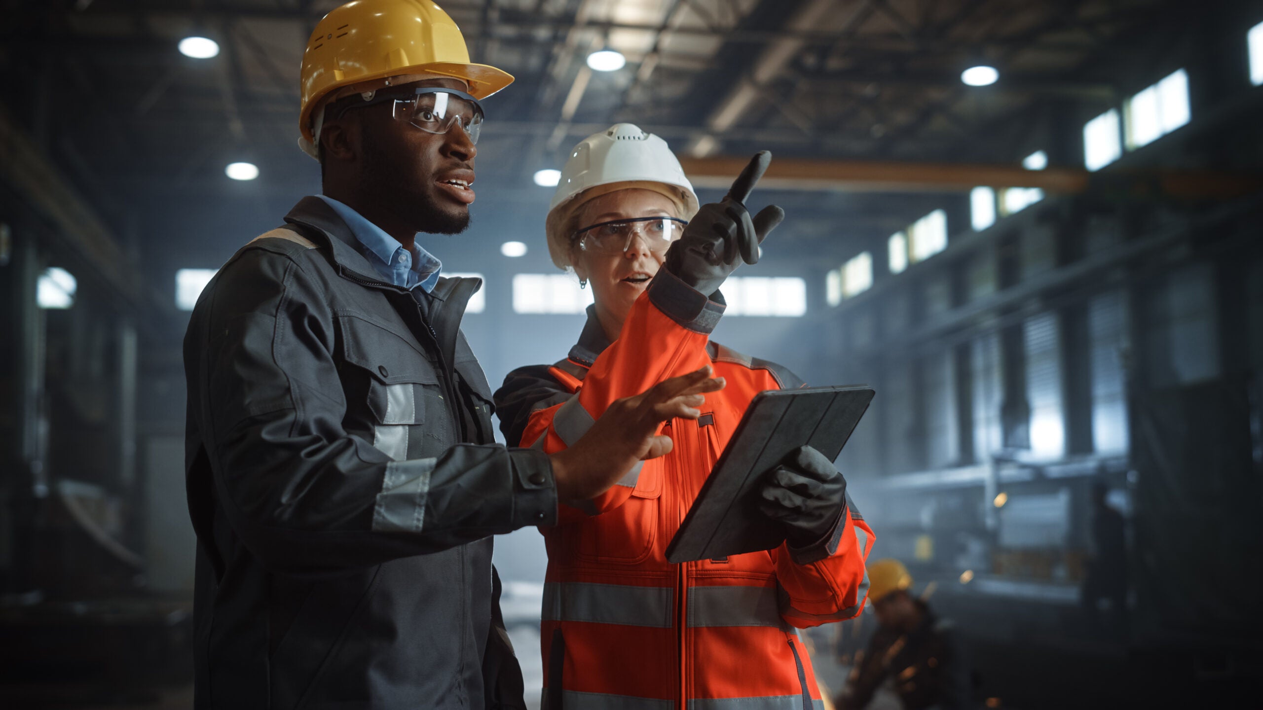 Two Heavy Industry Engineers Stand in Steel Metal Manufacturing Factory. One is holding a tablet and pointing, and the other is listening attentively