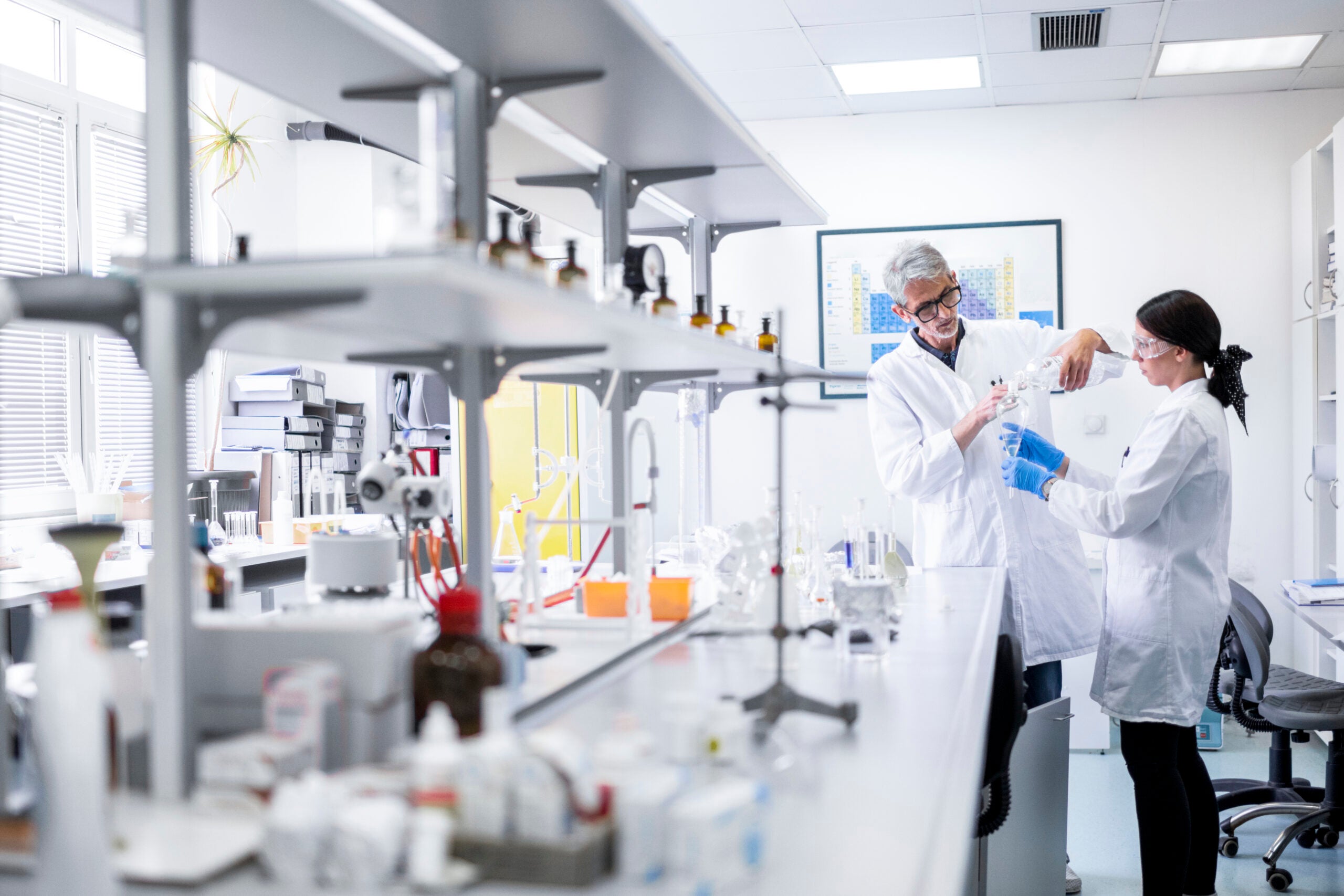 Two scientists in a laboratory, both wearing white lab coats and protective eyewear, stand at a lab counter with various scientific equipment and chemicals. The older scientist, with gray hair and glasses, is carefully pouring liquid from one glass container to another while instructing the younger scientist, who is wearing gloves and attentively watching. The lab is bright, with shelves filled with bottles and equipment, and a periodic table poster visible on the wall.