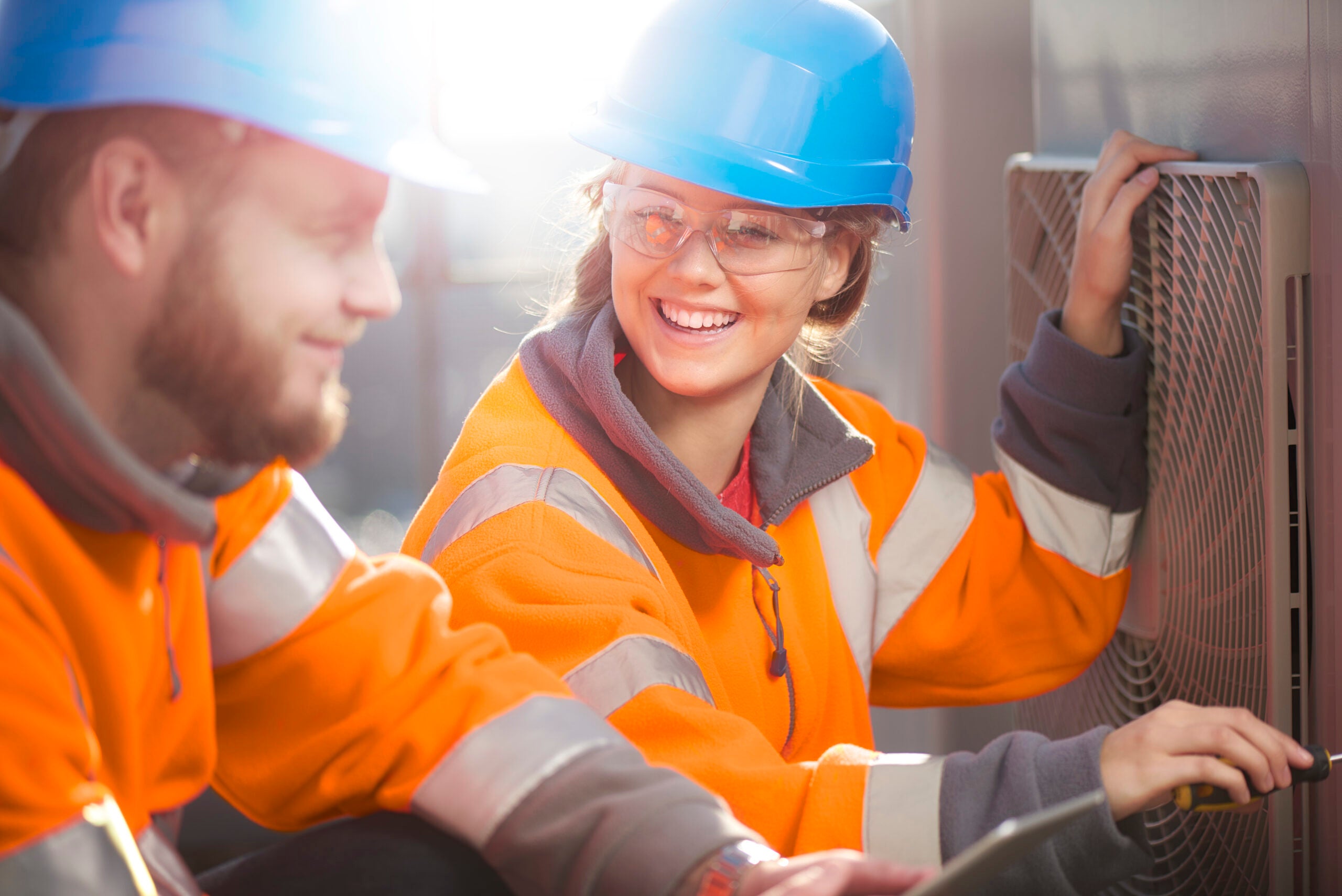 Two workers in high-visibility orange jackets and blue hard hats are performing maintenance or inspection work outdoors. The woman, smiling and holding a tool in one hand while steadying a large fan or vent with the other, appears engaged in conversation with her male coworker, who is looking at her and smiling. Both are wearing protective eyewear, and sunlight brightens the scene, adding a warm, positive atmosphere to their teamwork.