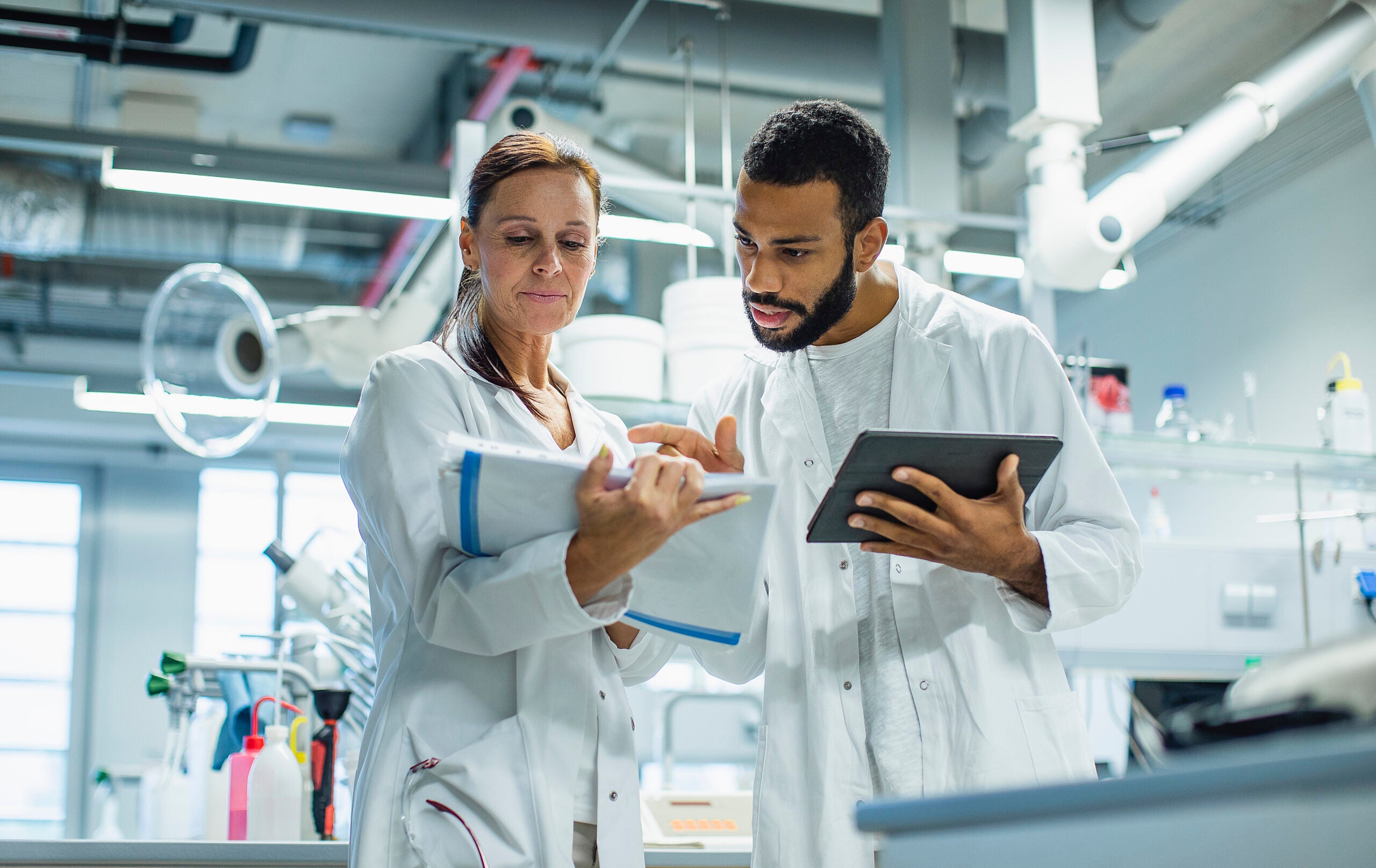 Two scientists in lab coats collaborate in a modern laboratory. The woman holds a clipboard, focusing on notes or data, while the man beside her holds a tablet, gesturing as he explains or discusses something. Both are engaged and serious, suggesting a professional exchange of ideas. The lab environment is filled with equipment, shelves, and bright overhead lighting, creating a high-tech and organized workspace.