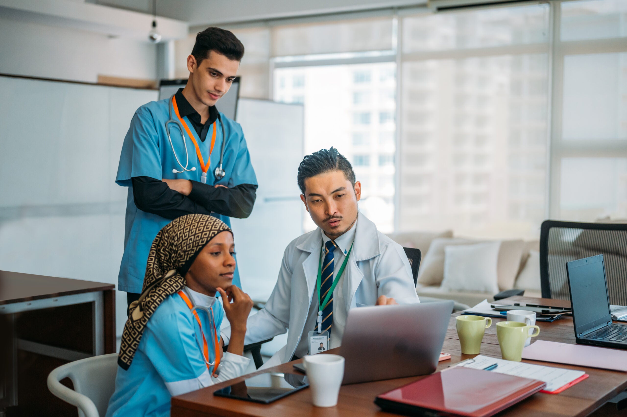 A diverse team of healthcare professionals, including two individuals in blue scrubs and one in a white lab coat, collaborate around a laptop in a bright, modern office. The doctor in the lab coat points at the laptop screen, explaining something to his colleagues, who listen attentively. One of the individuals in scrubs, a woman wearing a patterned headscarf, rests her chin on her hand, focused on the discussion. The workspace is organized with laptops, tablets, notebooks, and coffee cups, suggesting a professional yet comfortable environment for teamwork and learning.