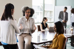 A group of women brainstorming together in a board room, with other businesspeople working independently in the background.