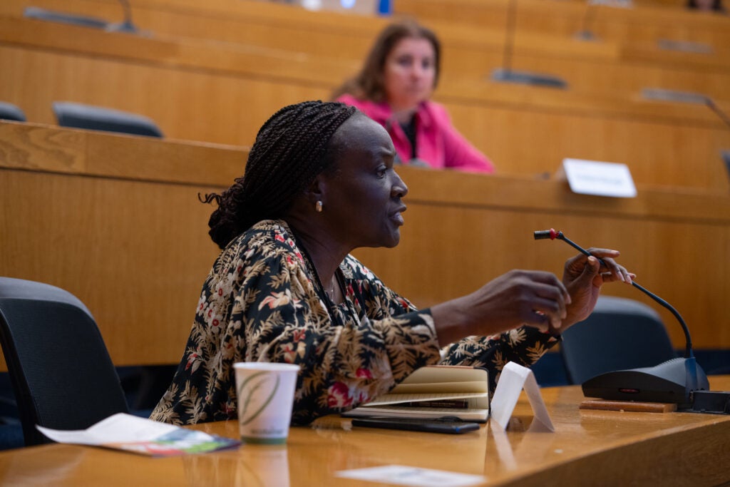 A woman with thin braids pulled into a low bun speaks earnestly into the microphone at hear seat in a lecture hall.