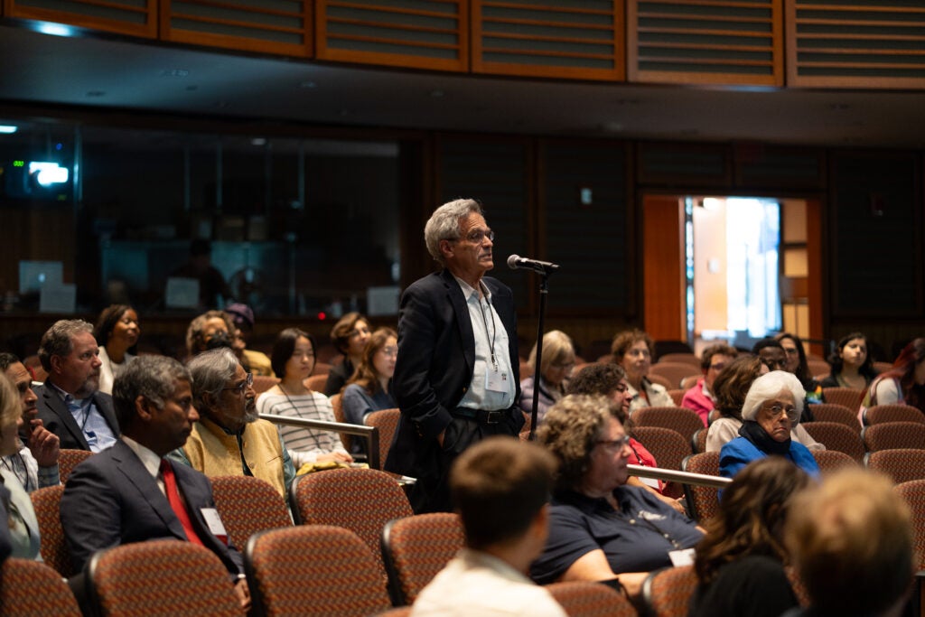 In the aisle of an auditorium, a man speaks into a standing mic as fellow audience members look on from their seats. 