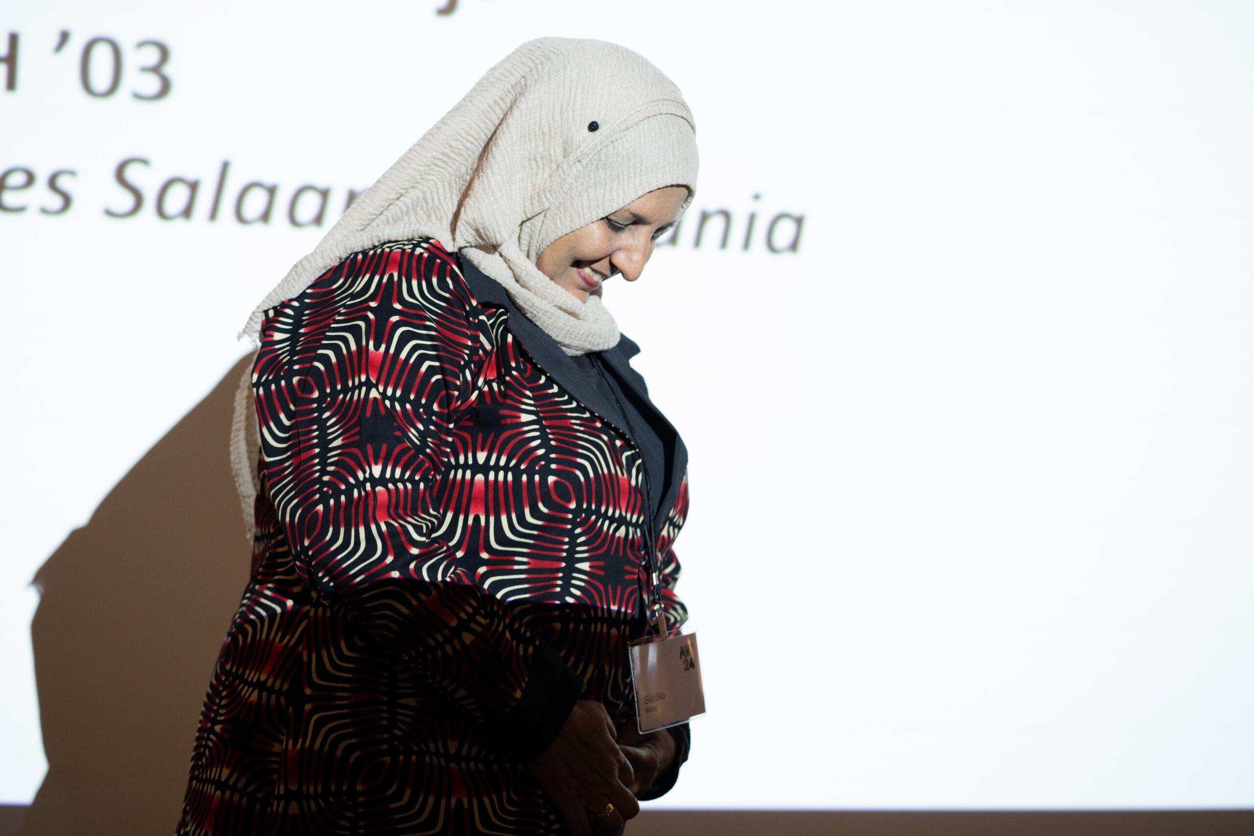 A woman wearing a white hijab and patterned red top smiles brightly with her head slightly bowed. She is standing in front of bright projector screen, appearing to listen to a speaker out of frame.