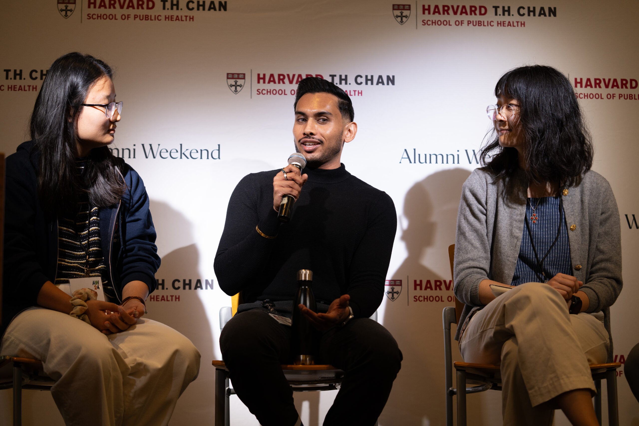 Three panel participants are seated in front of an Alumni Weekend background. In the center, a man dressed in all black speaks into a microphone. On either side, women with shoulder-length black hair and glasses turn their heads to listen.