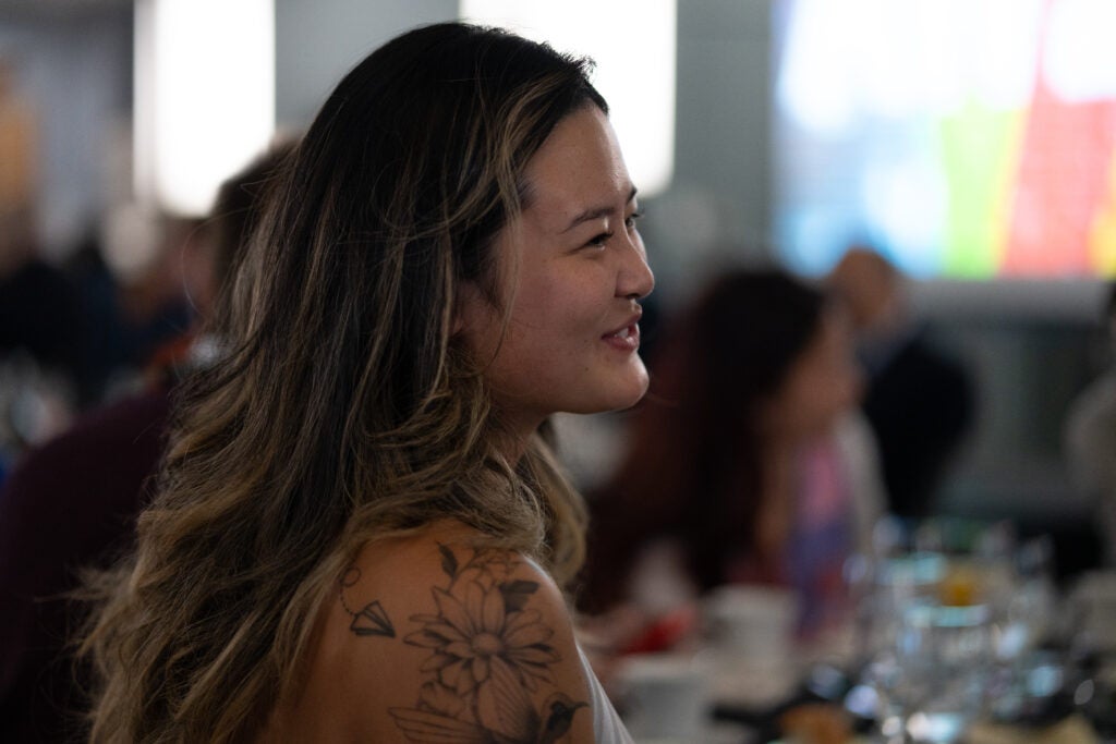 Shown in profile, a woman with highlighted dark hair and a floral tattoo on her shoulder smiles.