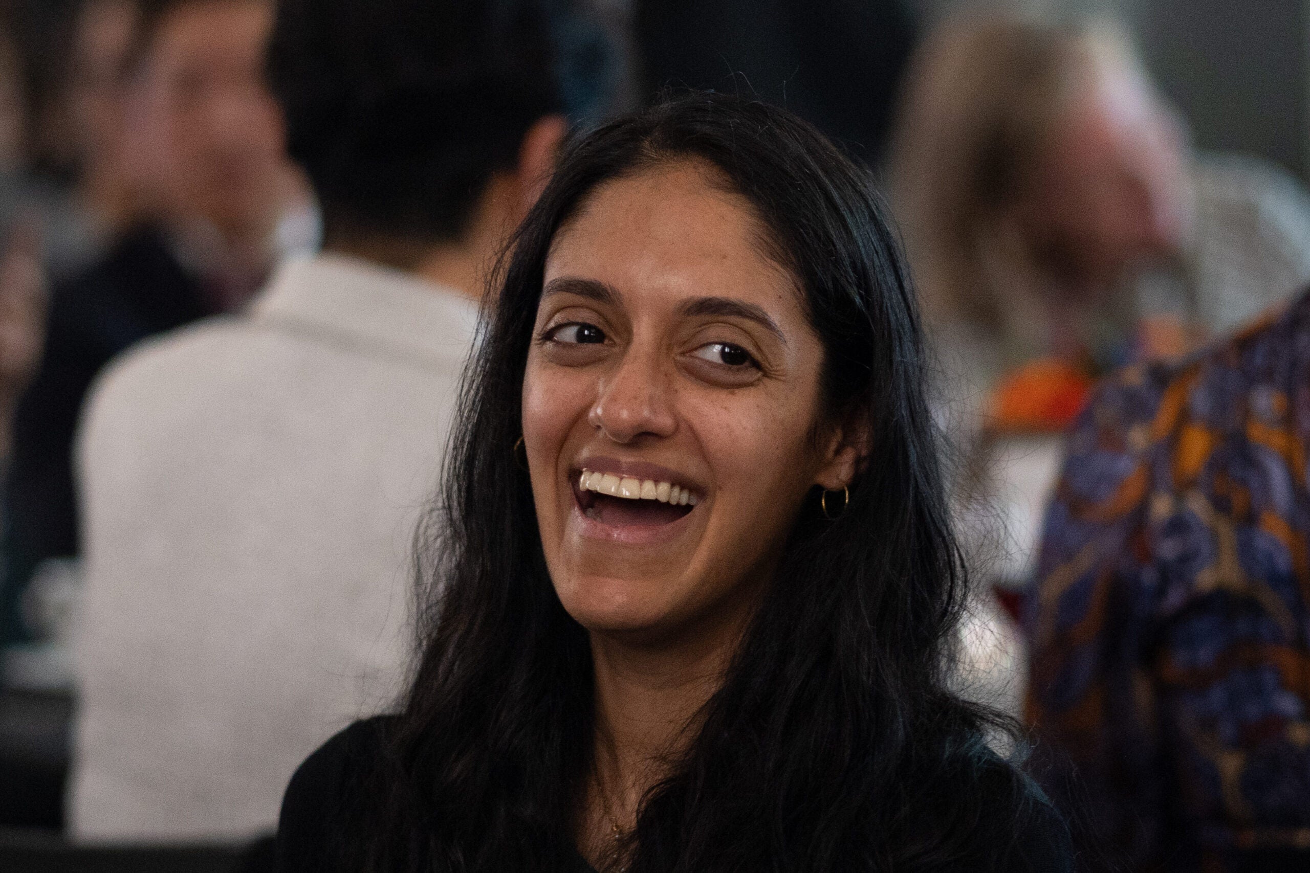 Seated at a table, a woman with long dark hair and small hoop earrings laughs heartily.