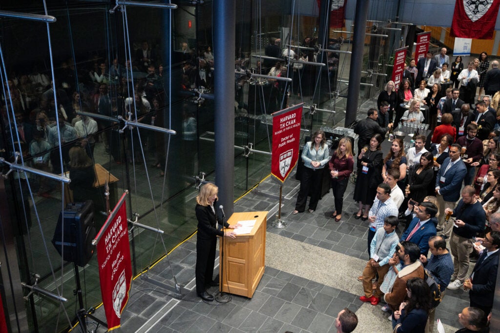 Alumni weekend attendees form a semicircle around a speaker at a podium in a glass atrium. The speaker wears a black suit and is flanked by banners that bear the School name and names of different degree programs.