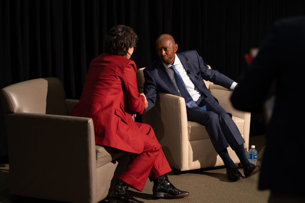 Two speakers seated in beige armchairs on stage shake hands. A Black man in a blue suit is facing the camera, and a short-haired woman in a red suit is facing him.