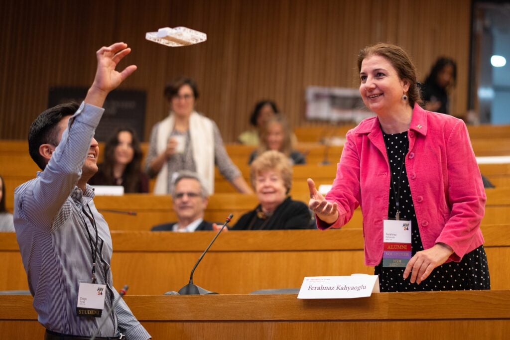 Standing in a lecture hall, a student in a blue striped shirt reaches up to catch an object thrown from the row behind him by an alumna Ferahnaz Kahyaoglu, who is wearing a pink corduroy jacket and black polka dot dress.