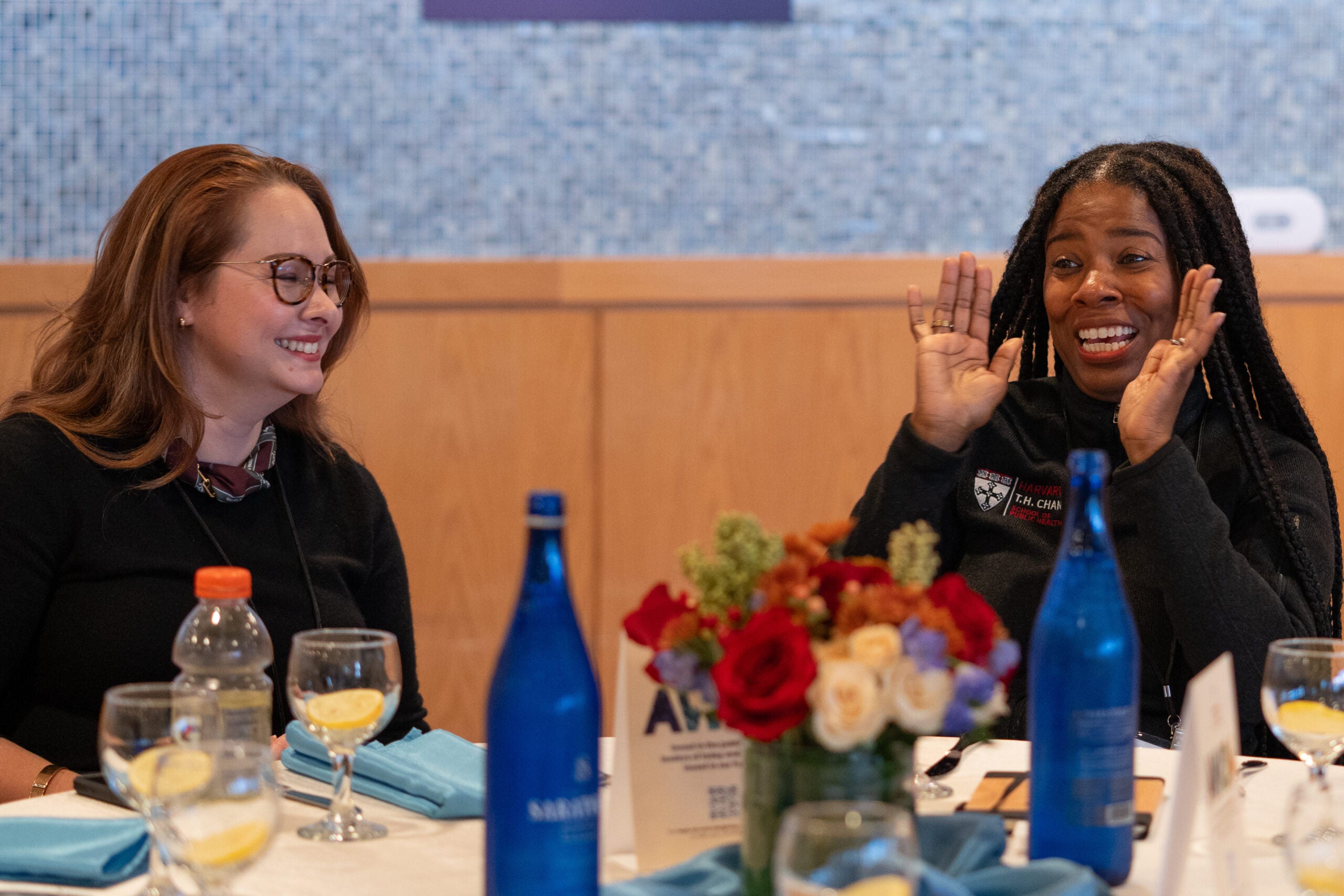 Seated at a set table, woman with medium brown hair and glasses laughs as a woman with long braids speaks with her hands up for emphasis.