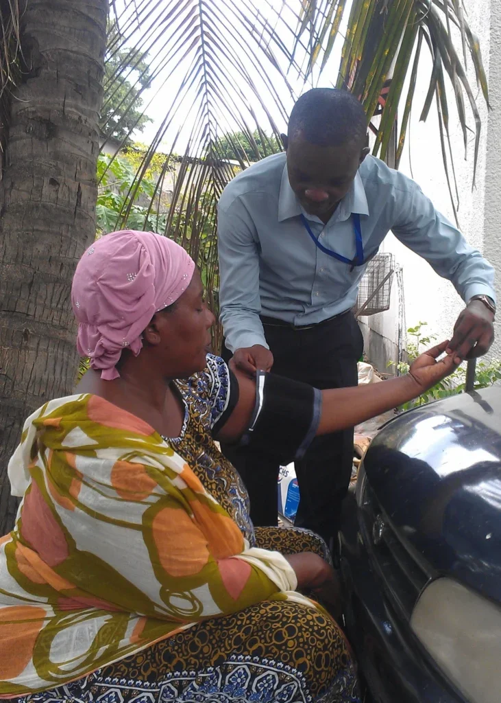 A medical professional checks a blood pressure cuff wrapped around a woman's arm. The woman is seated and wearing a pink head scarf. The pair are are outdoors, with palm trees in the background.