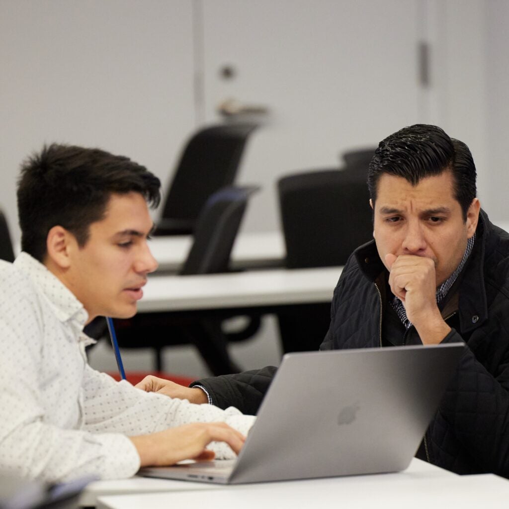 Two young men are looking at a laptop screen, appearing to work through a problem on it. One is speaking while the other has a focused look. 