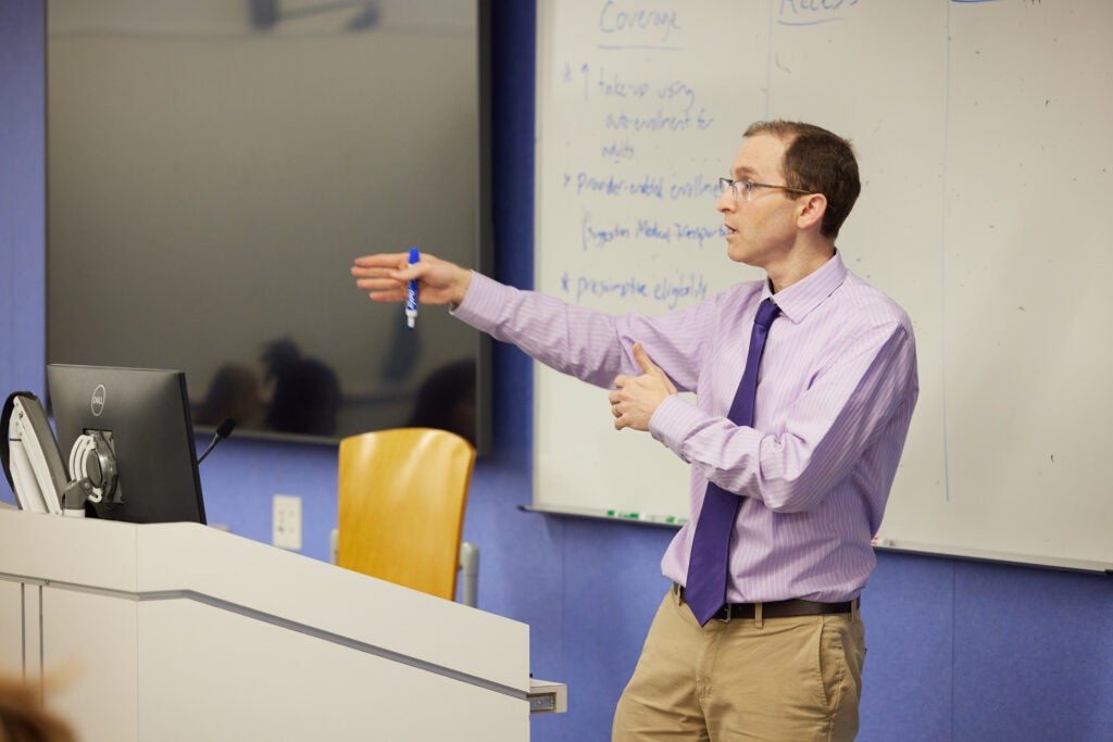 In a lavender shirt and purple tie, holding a dry erase marker, Ben Somers gestures while speaking behind a podium. 