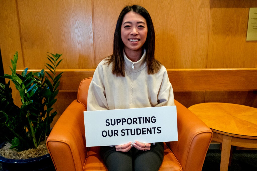 A young woman seated in the Kresge building atrium smiles, holding a sign that reads, "supporting our students."