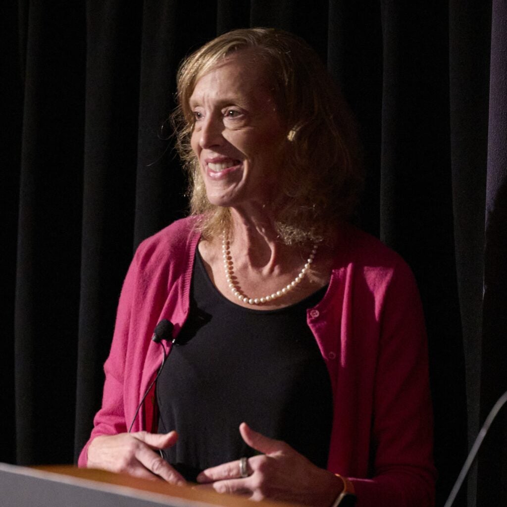 Sue Goldie is wearing a black top, pink sweater, and pearls, under stage lighting. 