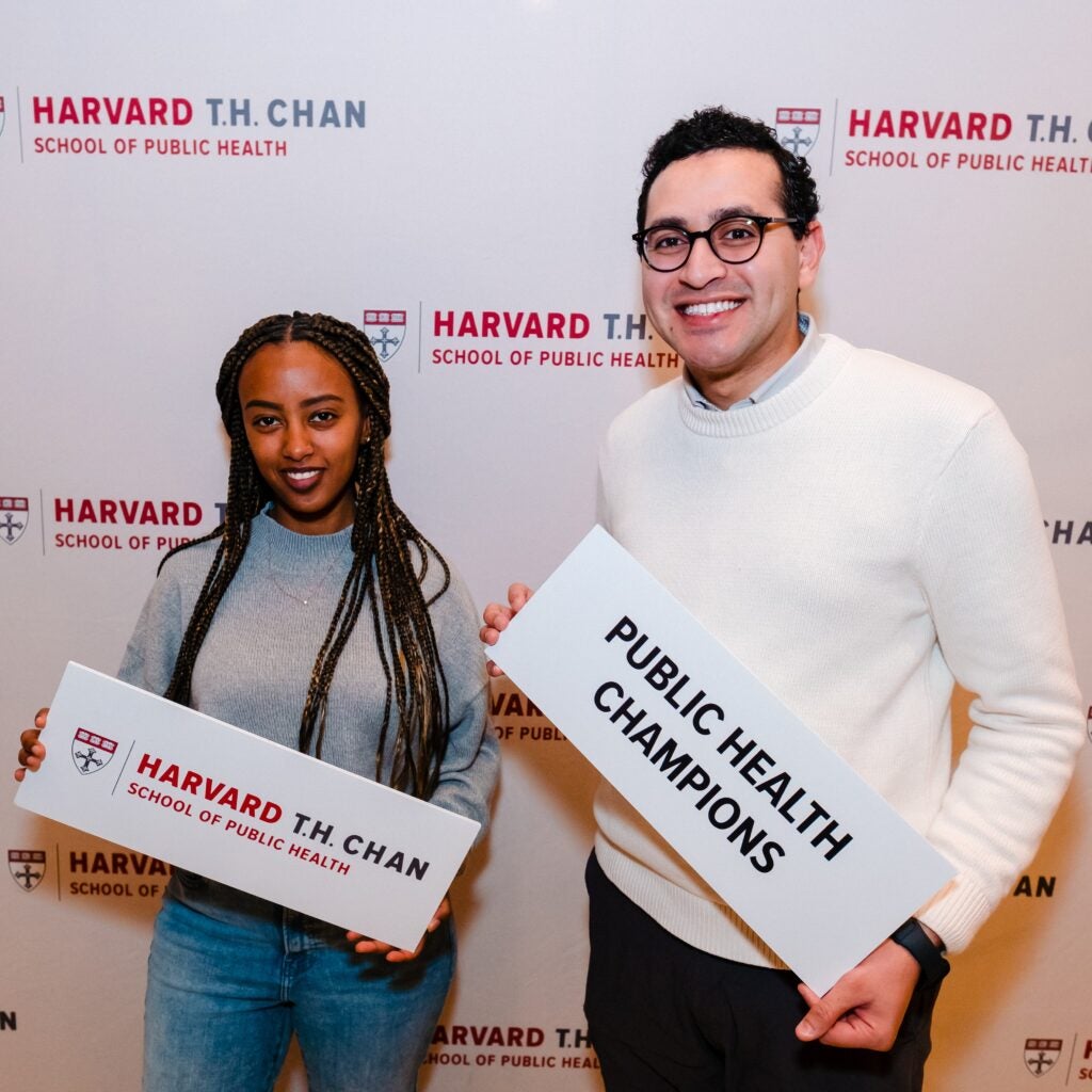 A woman with long braids a tall man with curly hair and glasses pose with signs in front of a white background with the school name and logo. The woman's sign is the School name and man's sign reads "Public Health Champions."