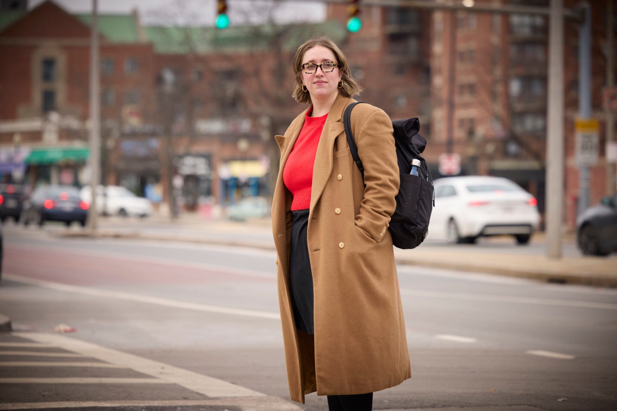 Jen Cuz stands at the edge of a city street in a long tan jacket, looking to her left