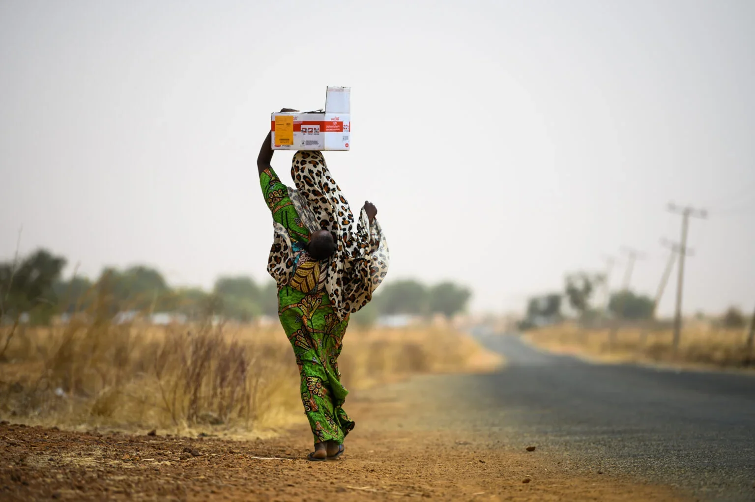 a mother carries her child on her back while carrying a box of food packets in Sokoto Nigeria