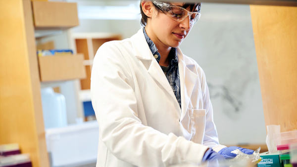 Lab member wearing a white coat and goggles working in the lab.