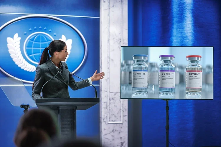 A woman standing behind a podium speaking to a crowd and gesturing to a screen with images of the COVID-19 vaccine