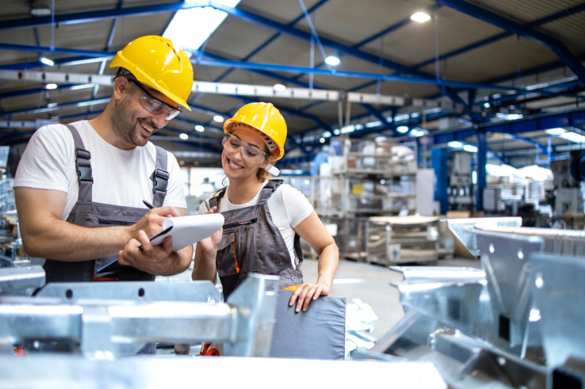 Two smiling workers wearing PPE standing in a factory, looking over notes on a clipboard