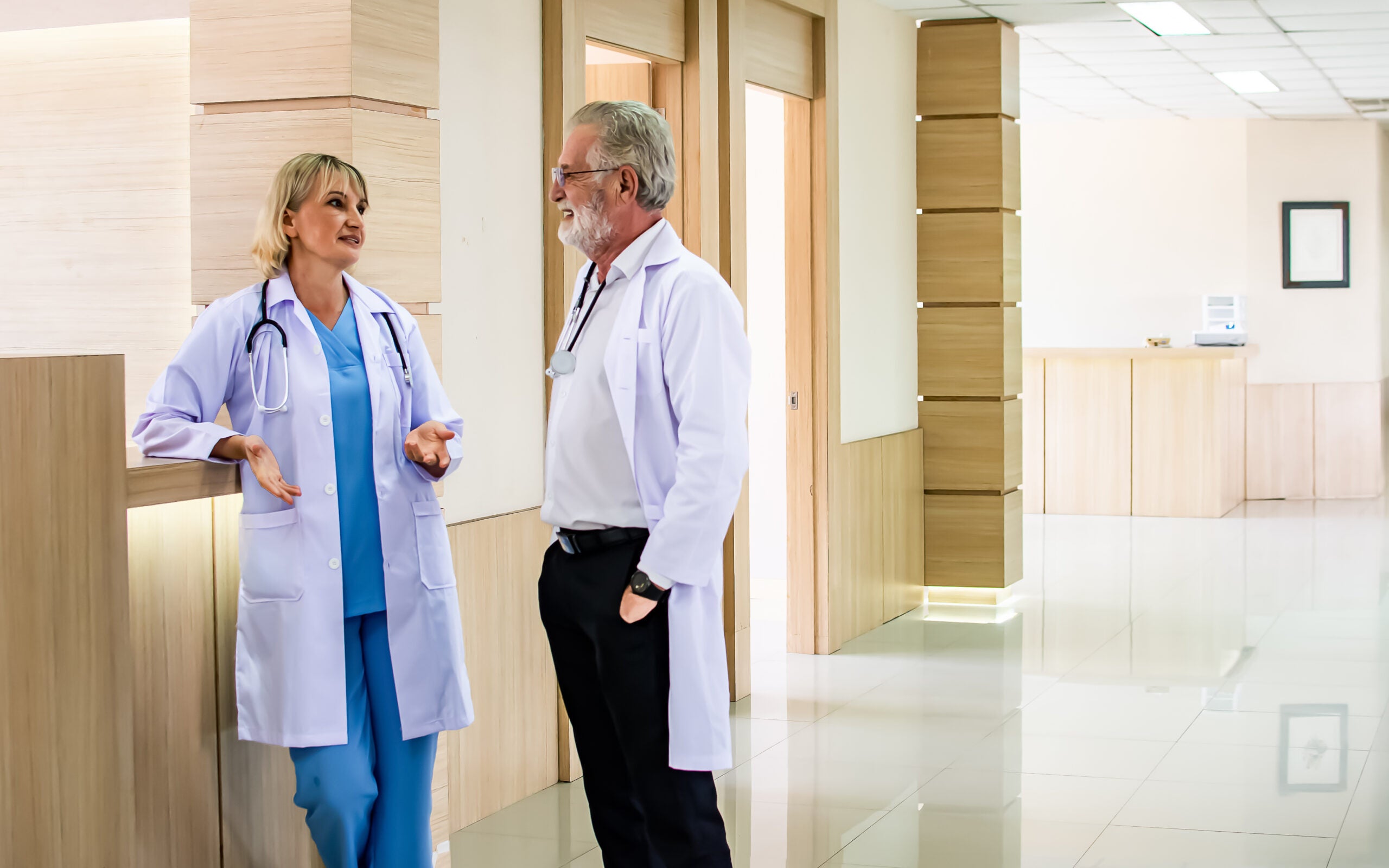 A male and female doctor standing by the reception desk in a hospital, talking.