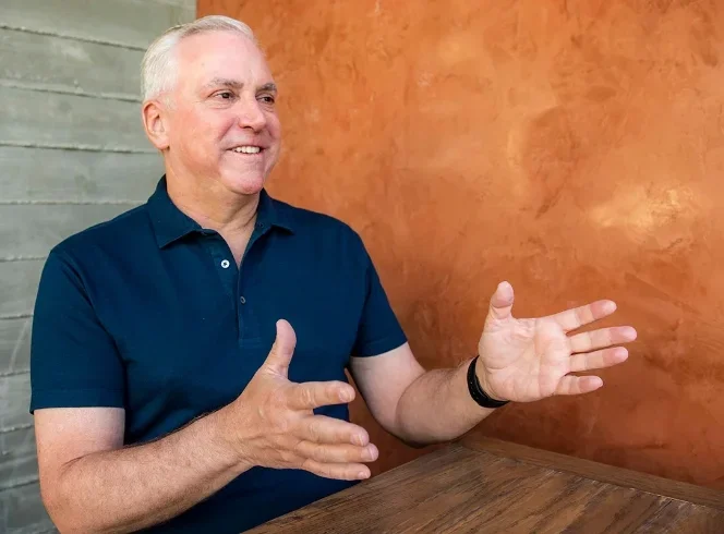 Michael Dillon, seated at a table and wearing a navy blue polo shirt, gesturing as a smiles. 