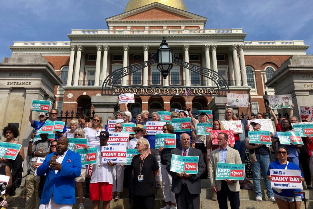 A group of protestors in front of the Masscushetts state house. They hold signs that read "Today is a rainy day governor!" and "Keep carney hospital open".