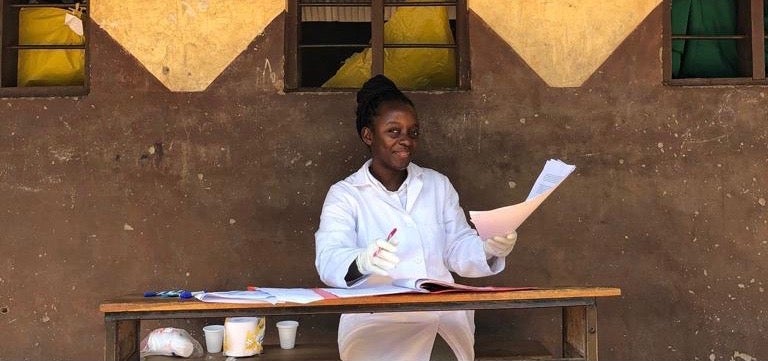 Valencia Lambert sits at a desk, in a white coat and gloves, in front of an earth-toned wall. She is handling papers and smiling.