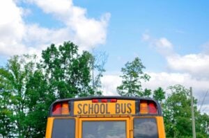 Photo of a yellow school bus in front of a blue sky and green trees.