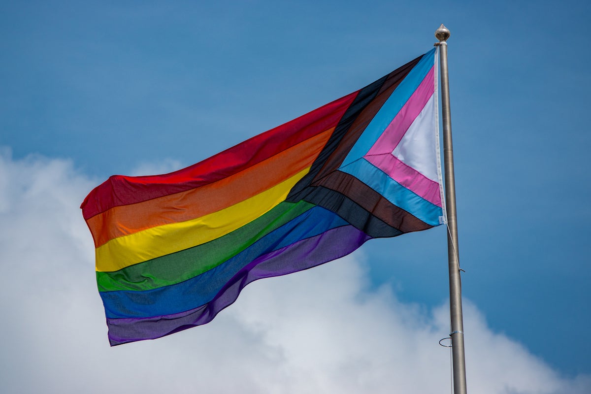 a progress pride flag flying in the wind against a blue sky