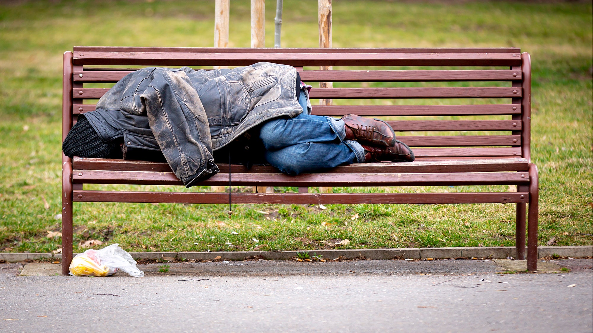 Homeless man sleeps on a bench covered with his jacket in a public park during the day