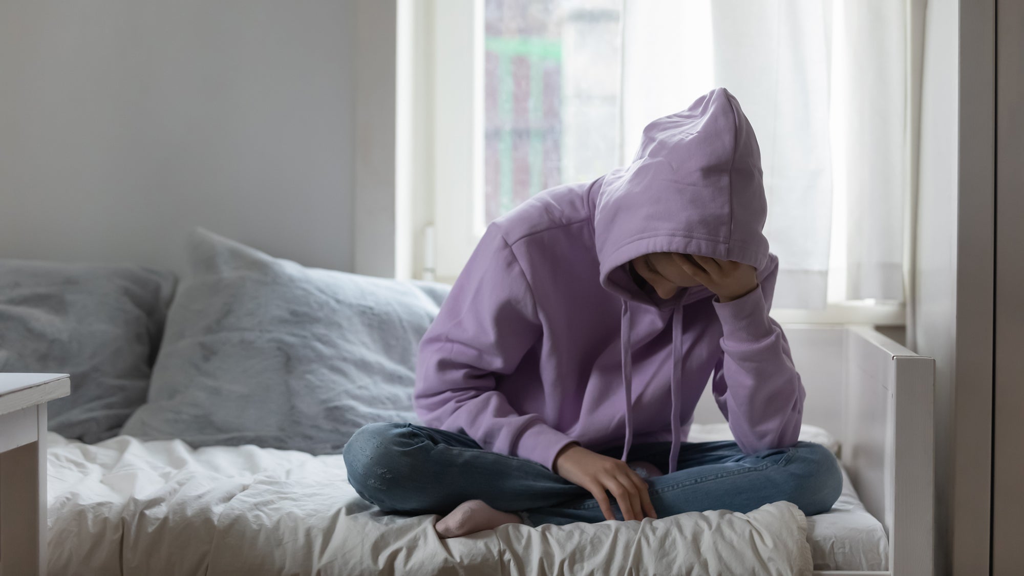 A teen in a hoodie sitting on their bed, holding their head