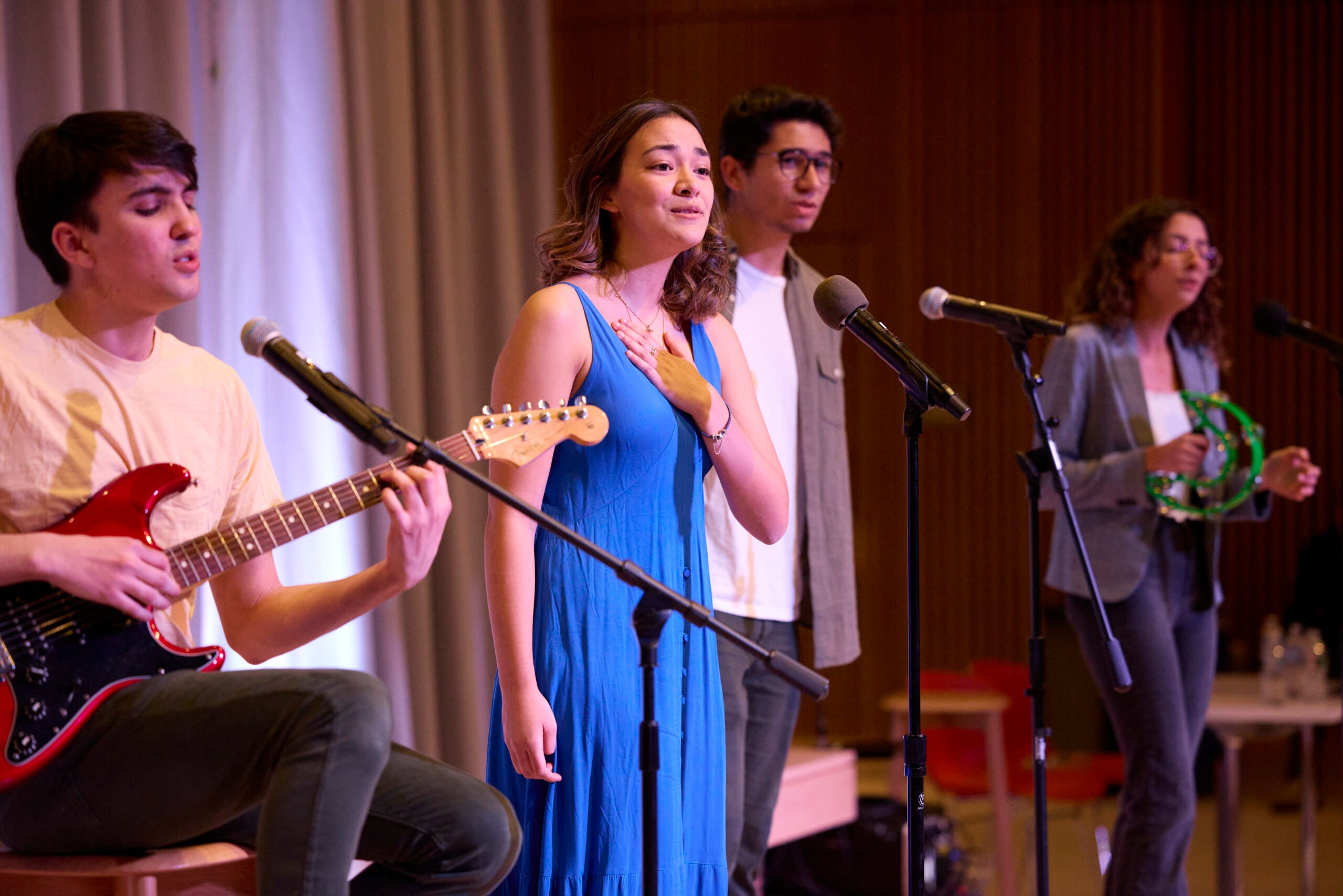 A person in a blue dress sings with a hand over her heart. She's surrounded by other people holding musical instruments and singing.