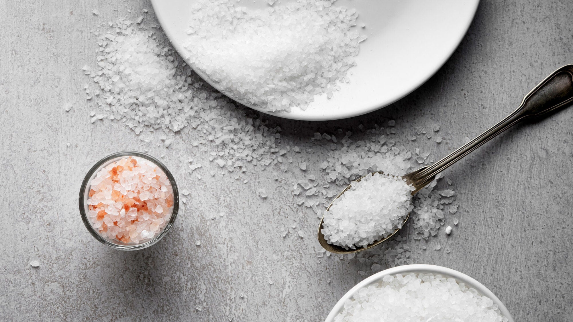 White salt in small bowls and a spoon spill onto a gray surface. A small bowl of pink sea salt is on the left.