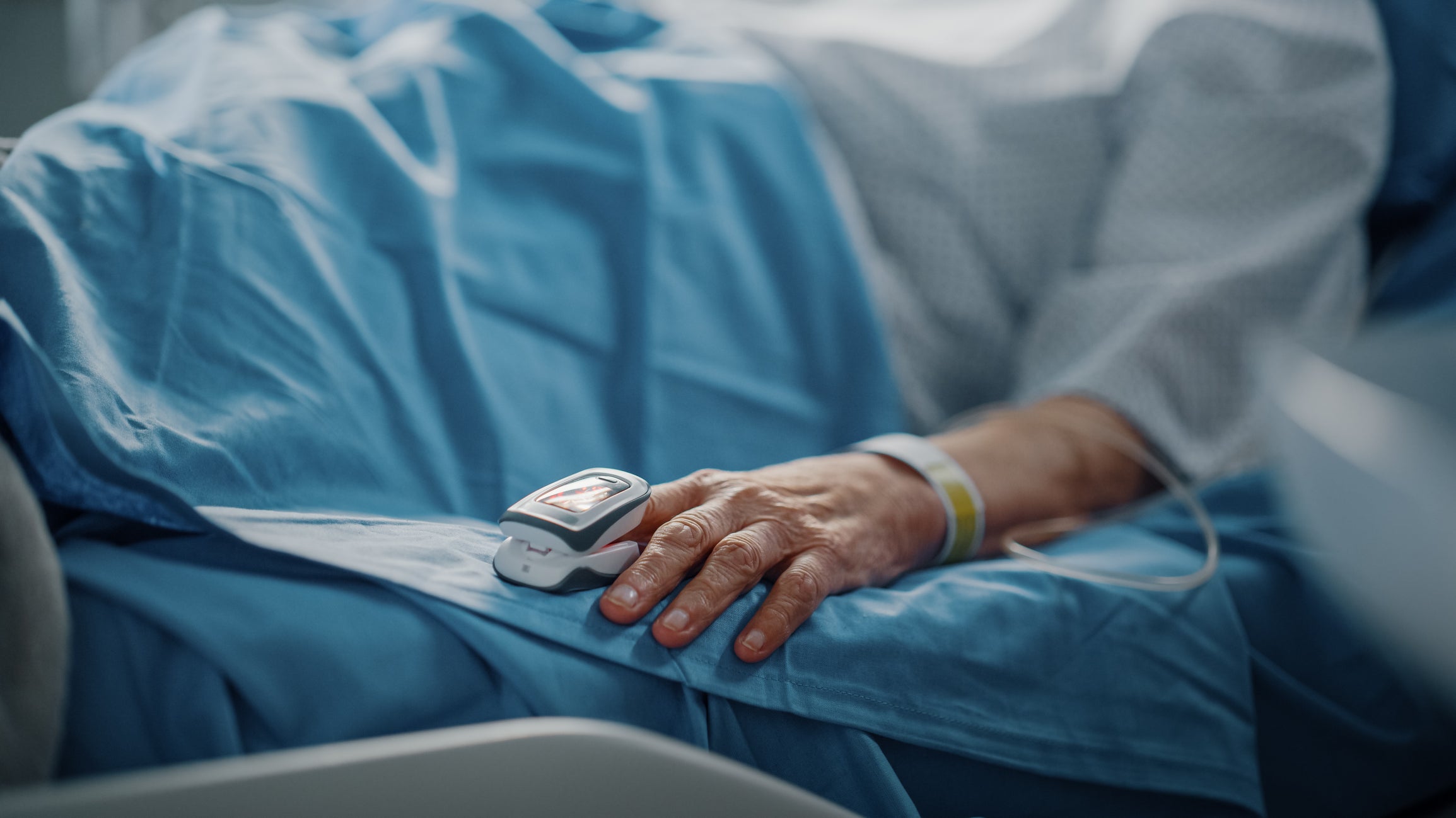 Hospital Ward: Senior Woman Resting in a bed with Finger Heart Rate Monitor / Pulse oximeter showing Pulse. Her Fragile Hands Resting on a Blanket. Focus on the Hand.