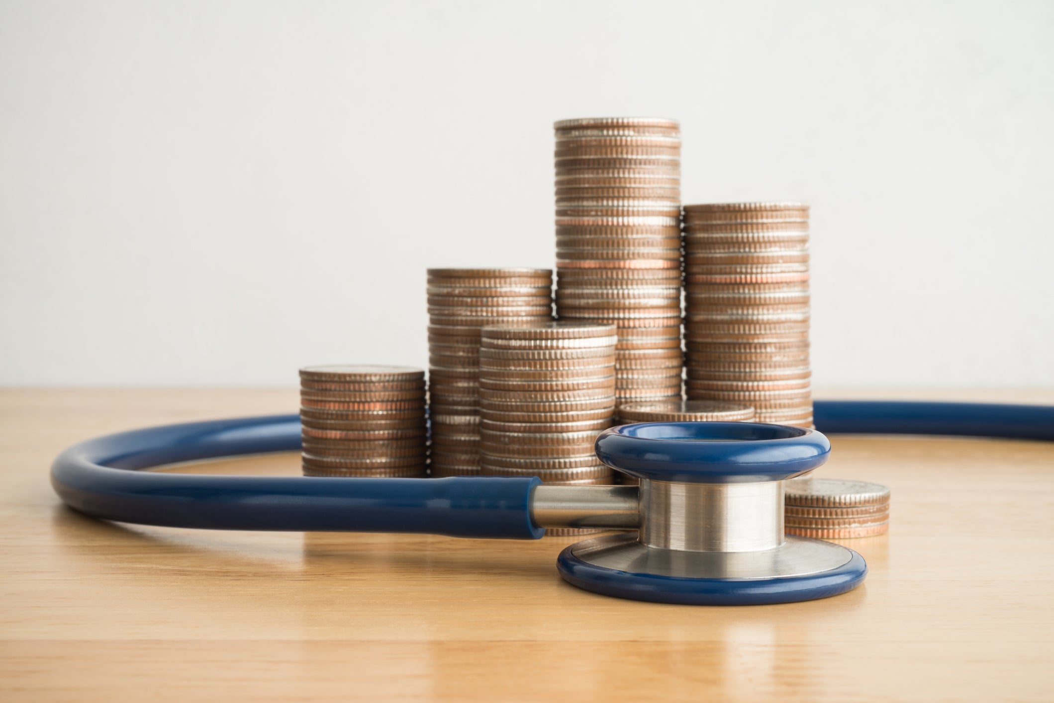 Stethoscope wrapped around stacked coins on wooden table. white wall background.