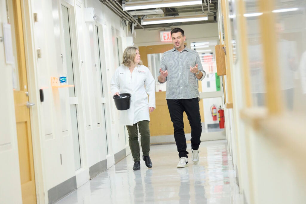 Will Mair, in a grey button-down shirt, black pants, and sneakers, walks through a hallway with a colleague in a white lab coat, black pants and black shoes.