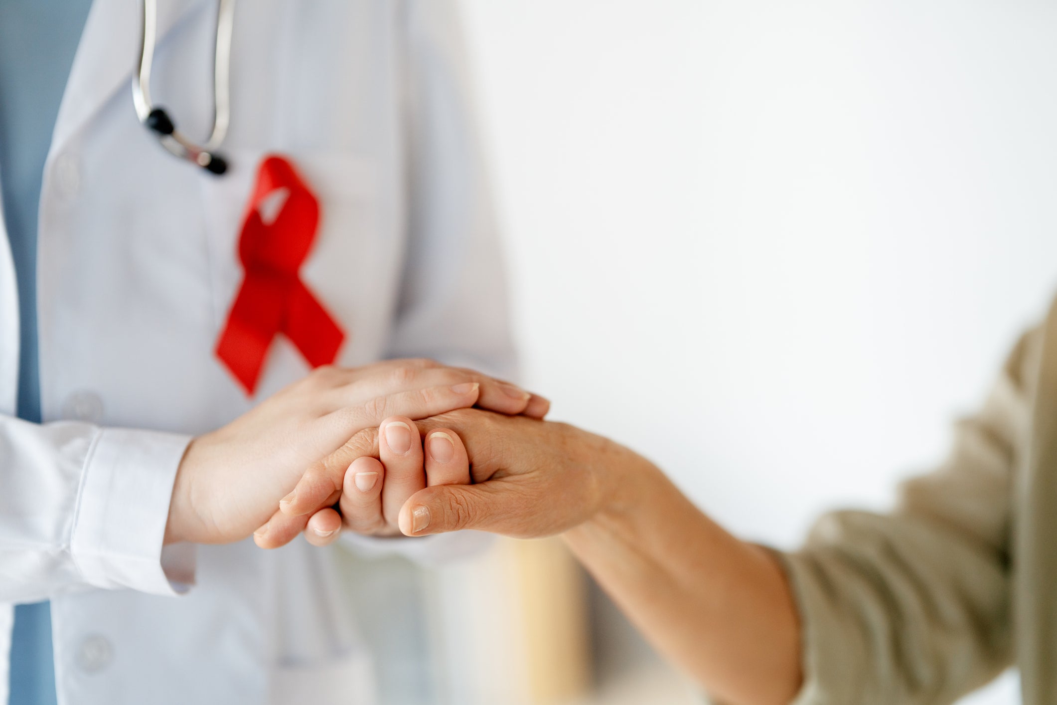 Doctor wearing red ribbon, symbolizing support for people living with HIV/AIDS, holds the hand of a patient. Image excludes faces, close-up of hands.
