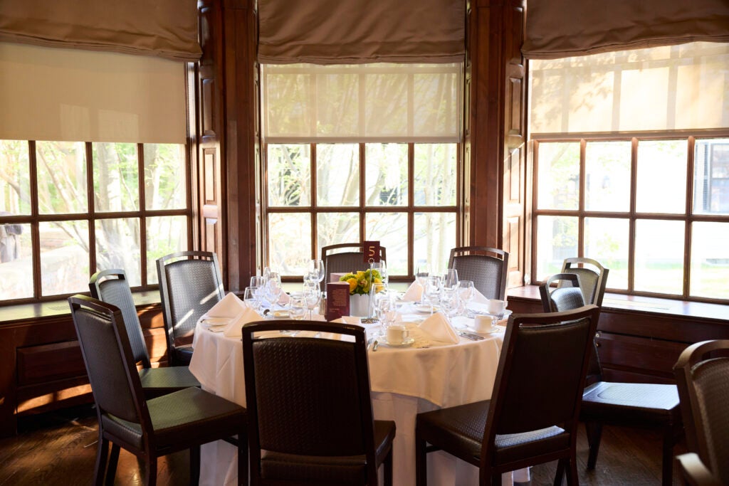 A round table set for eight stands ready in front of wood-paneled windows with the shades partially drawn. 