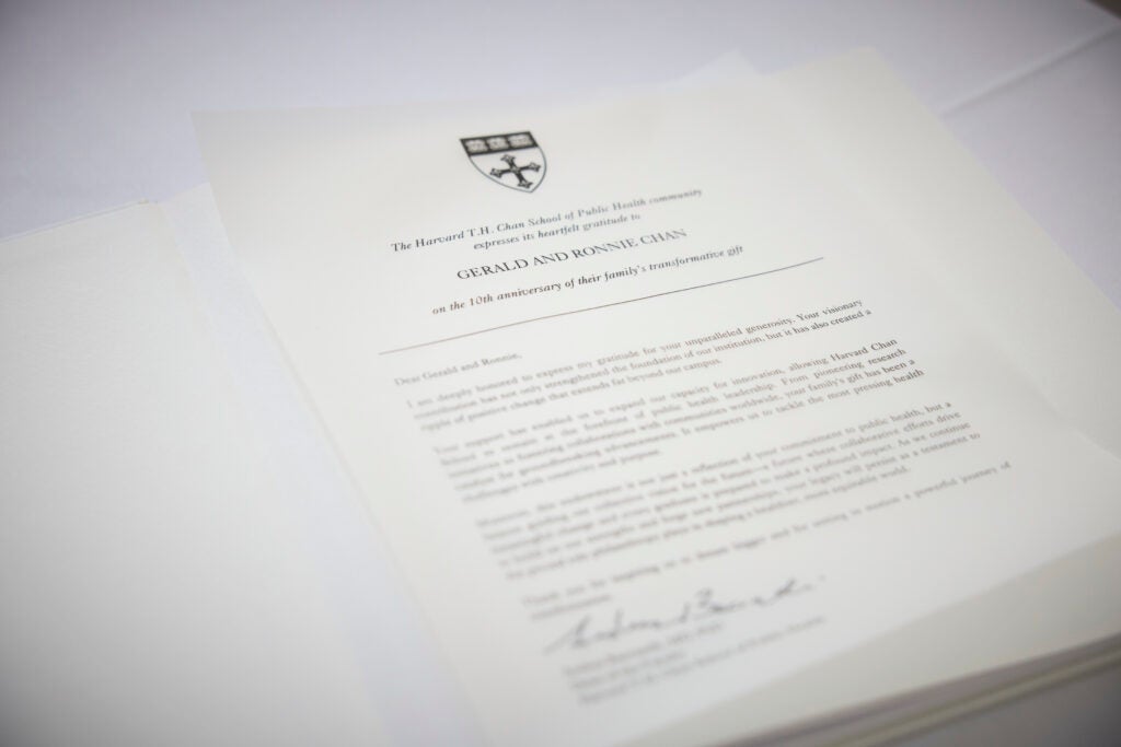 A document bearing the School crest reads in Large font at the top: The Harvard T.H. Chan School of Public Health Community expresses its heartfelt gratitude to Gerald and Ronnie Chan on the 10th anniversary of their family's transformative gift. The rest of the letter is blurred, but Dean Baccarelli's signature can be seen at the bottom. 