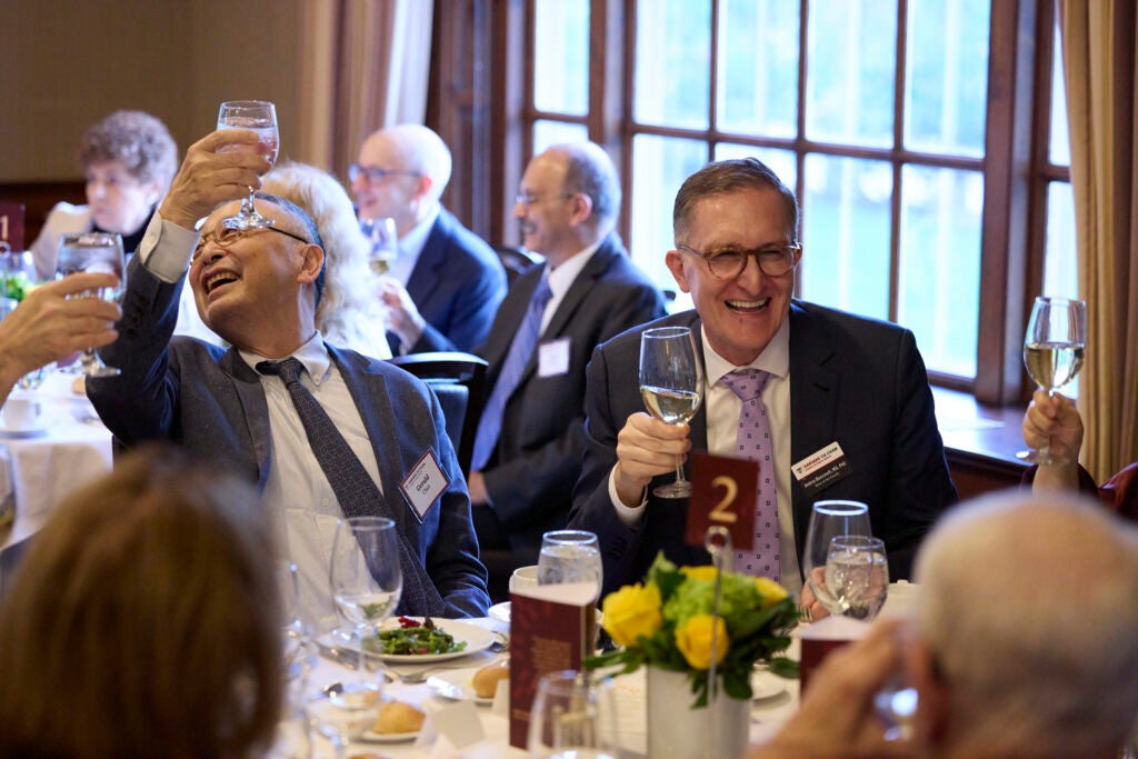 Gerald Chan and Dean Baccarelli are seated next to each other at table 2. Both are smiling with their glasses raised in a toast, and Gerald is looking over his raised arm at someone out of frame. 