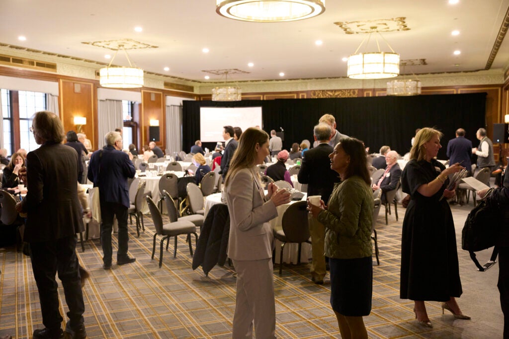 The conference room is full of guests engaged conversing at their tables or in small standing groups. 