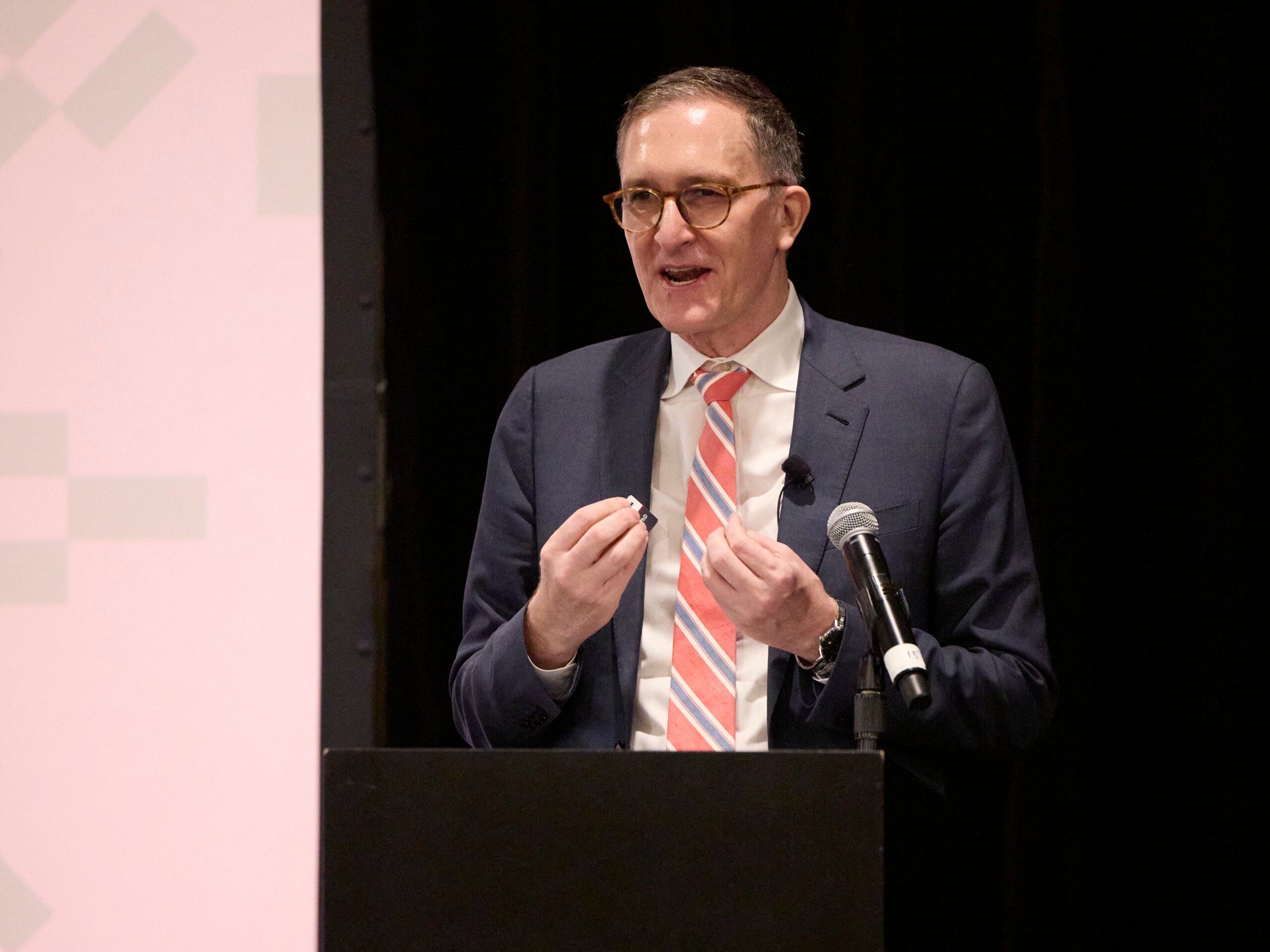 The Dean speaks at the podium, wearing a dark blue jacket, white shirt, and tie with red and gray stripes